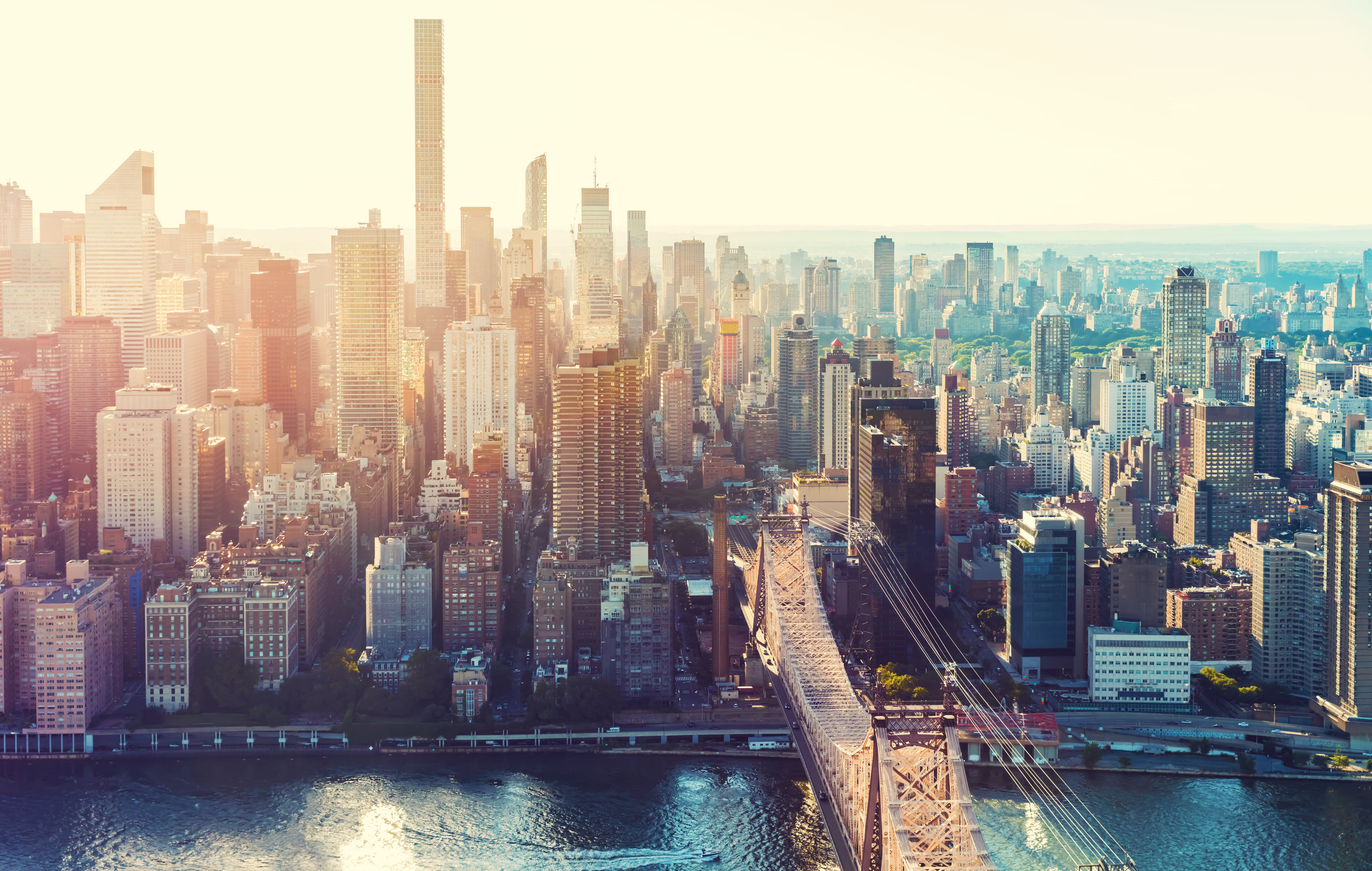 Aerial view of the New York City skyline featuring the Queensboro Bridge over water, illuminated by golden hour light.