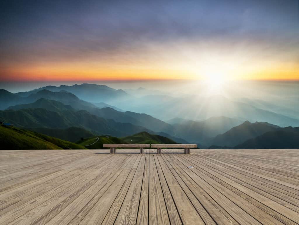 A wooden deck with two benches overlooking a panoramic mountain range at sunrise or sunset, with a vibrant sky and sunburst.