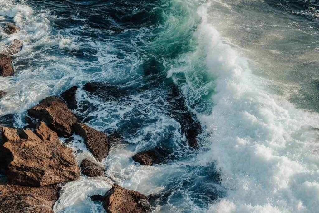 Overhead view of powerful ocean waves, with white foam and spray, crashing against dark, rugged rocks.