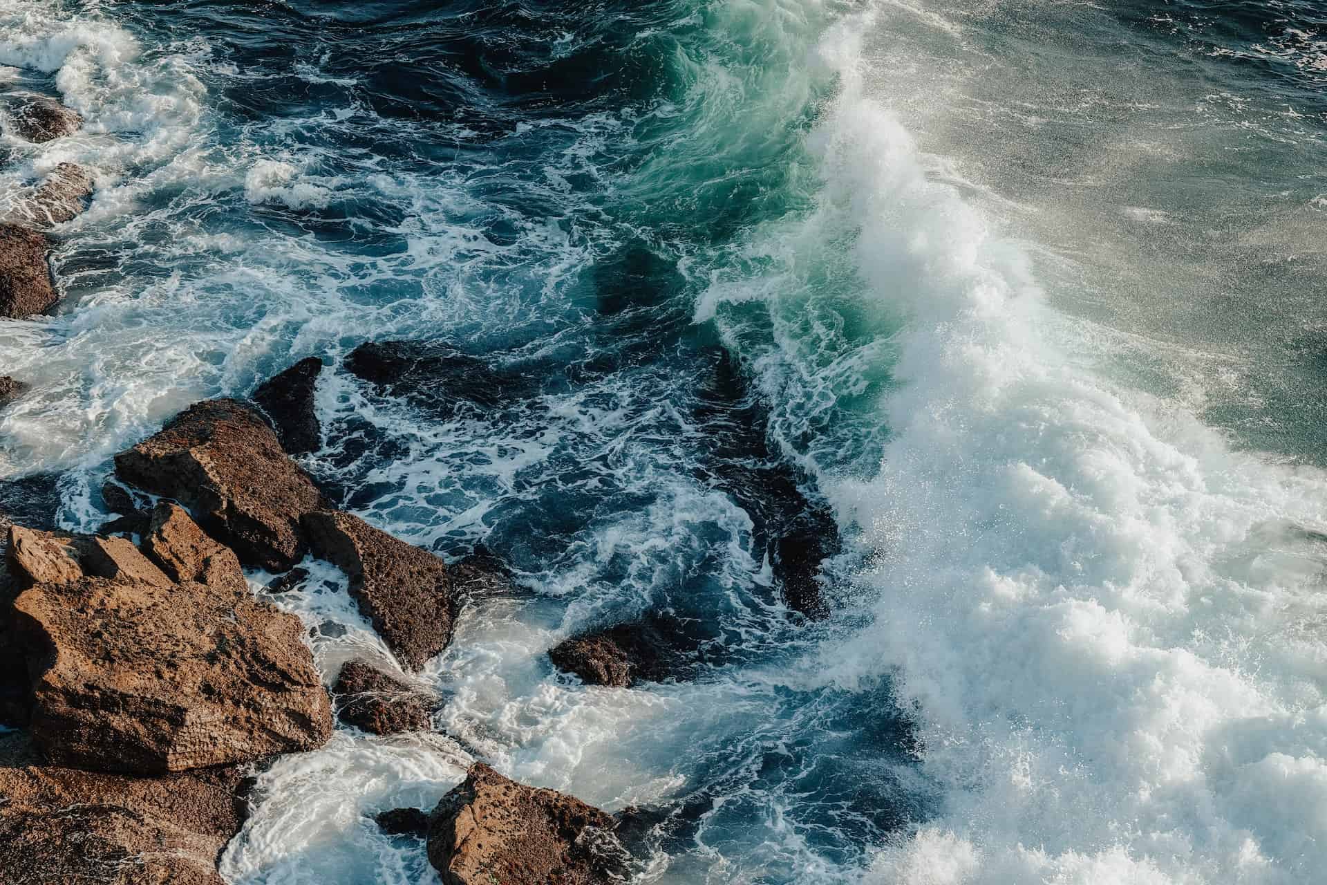 Overhead view of powerful ocean waves, with white foam and spray, crashing against dark, rugged rocks.