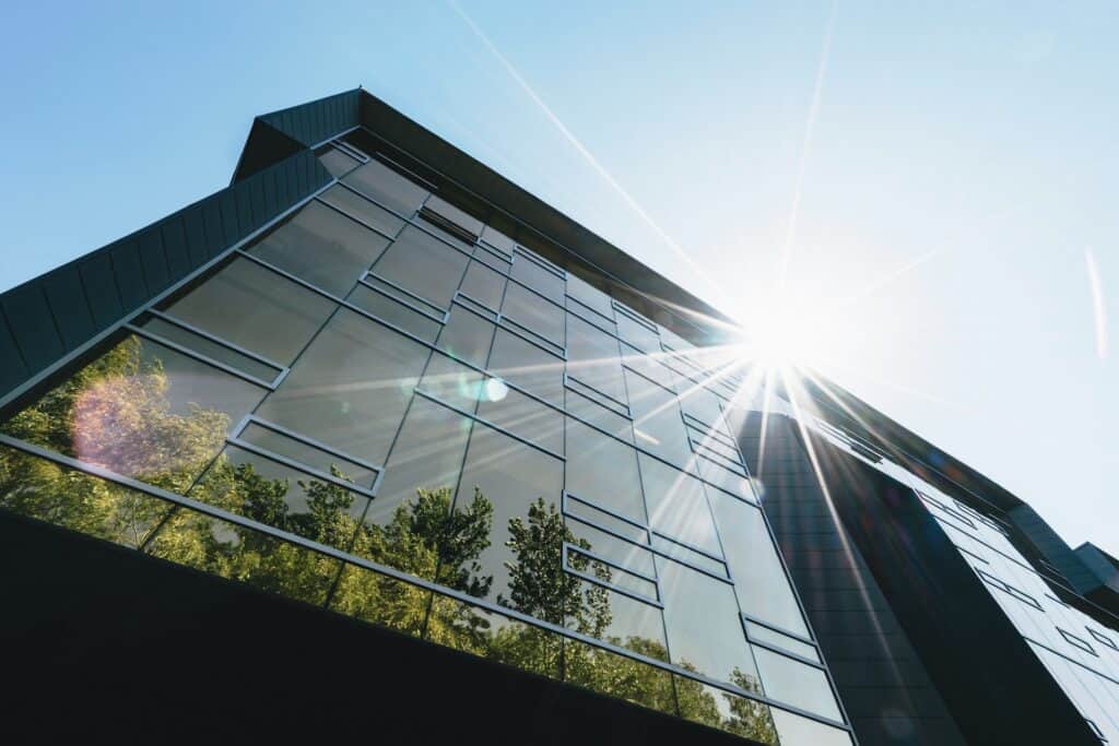 Low-angle view of a modern glass building reflecting green trees, with a bright sunburst in the clear blue sky.