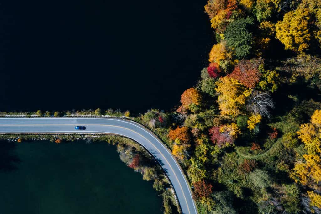 Aerial view of a winding road with a blue car, separating two dark bodies of water, surrounded by colorful autumn trees.