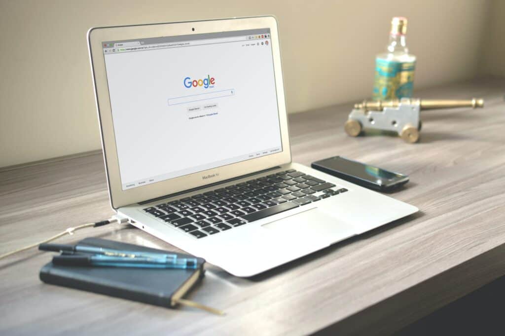 A silver MacBook Air laptop on a wooden desk, displaying the Google search page. A notebook, pens, a smartphone, a miniature cannon, and a bottle are also on the desk.