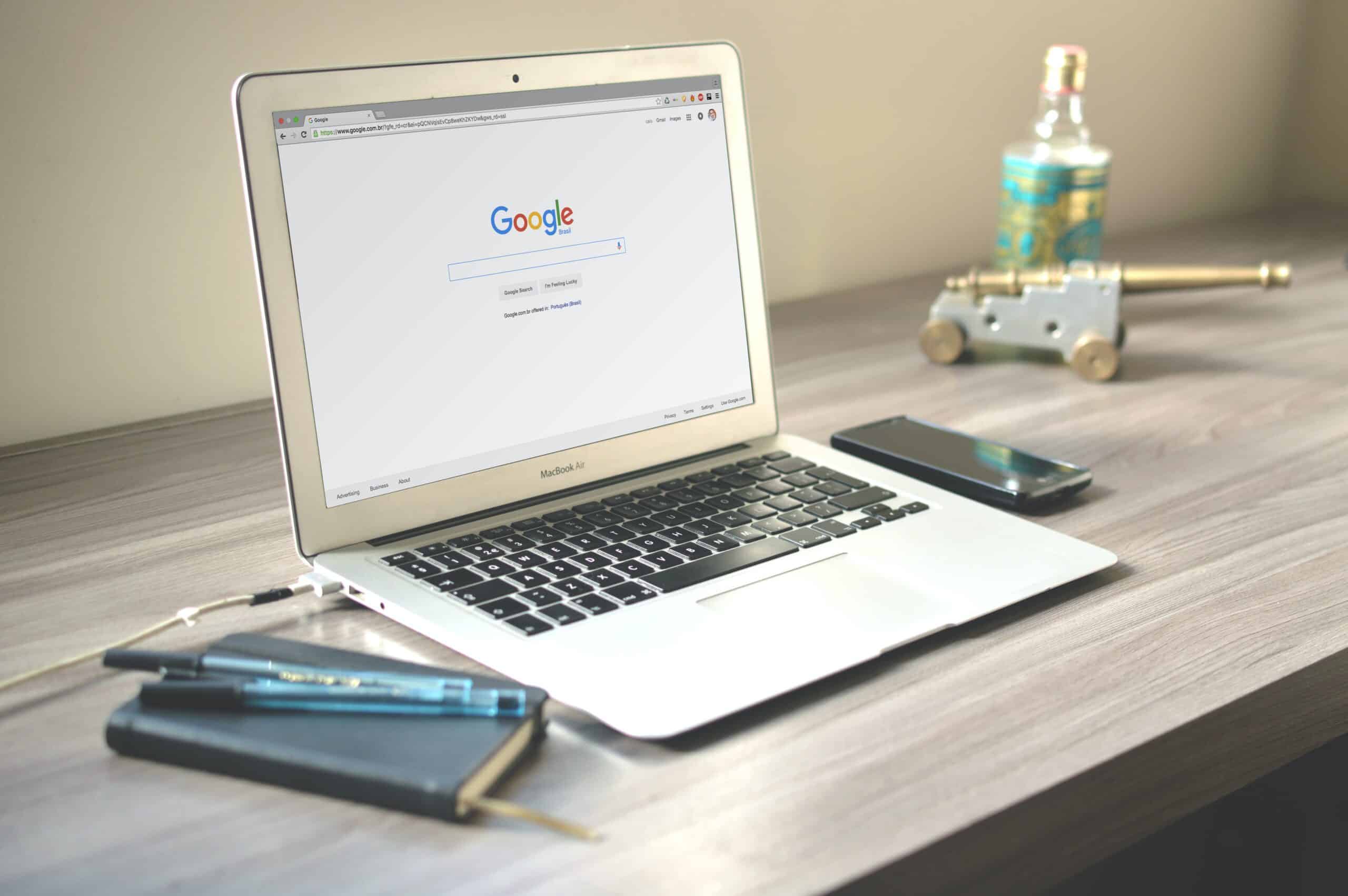 A silver MacBook Air laptop on a wooden desk, displaying the Google search page. A notebook, pens, a smartphone, a miniature cannon, and a bottle are also on the desk.