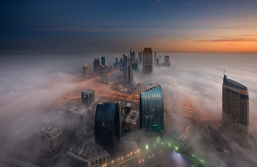 Aerial view of a city skyline at dawn or dusk, with skyscrapers rising above a thick layer of fog and illuminated roads visible below.