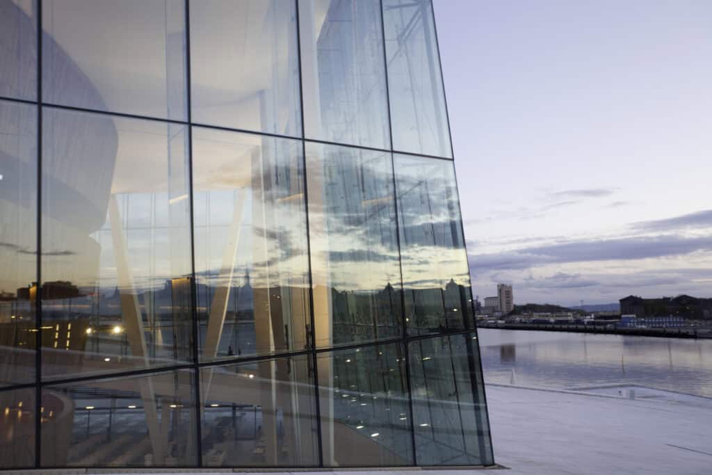 A modern glass building with reflections of the sky, clouds, and a distant city across a body of water at dusk.
