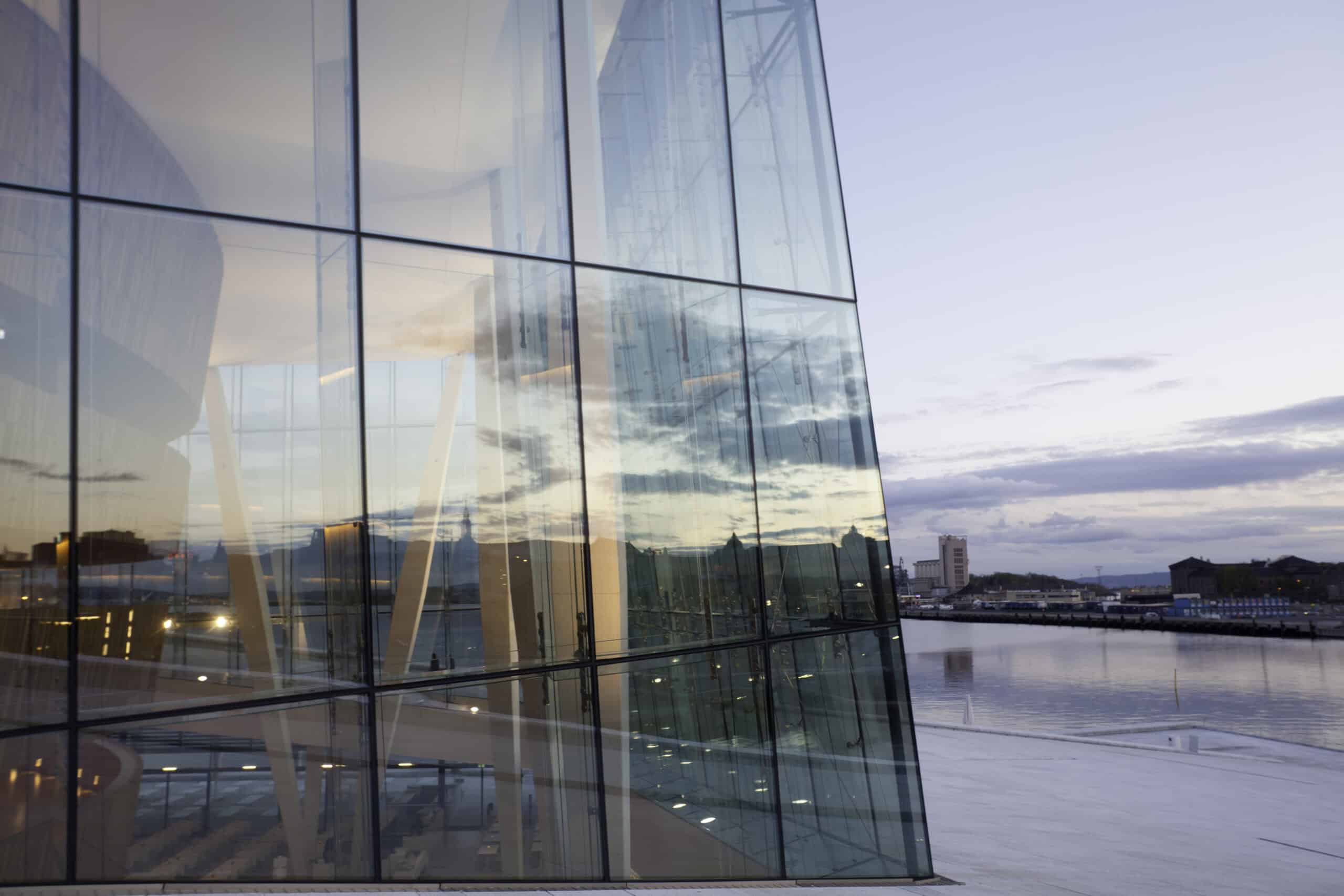 A modern glass building with reflections of the sky, clouds, and a distant city across a body of water at dusk.
