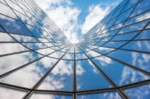 A modern glass building reflecting a blue sky with white clouds, viewed from a low angle looking up.