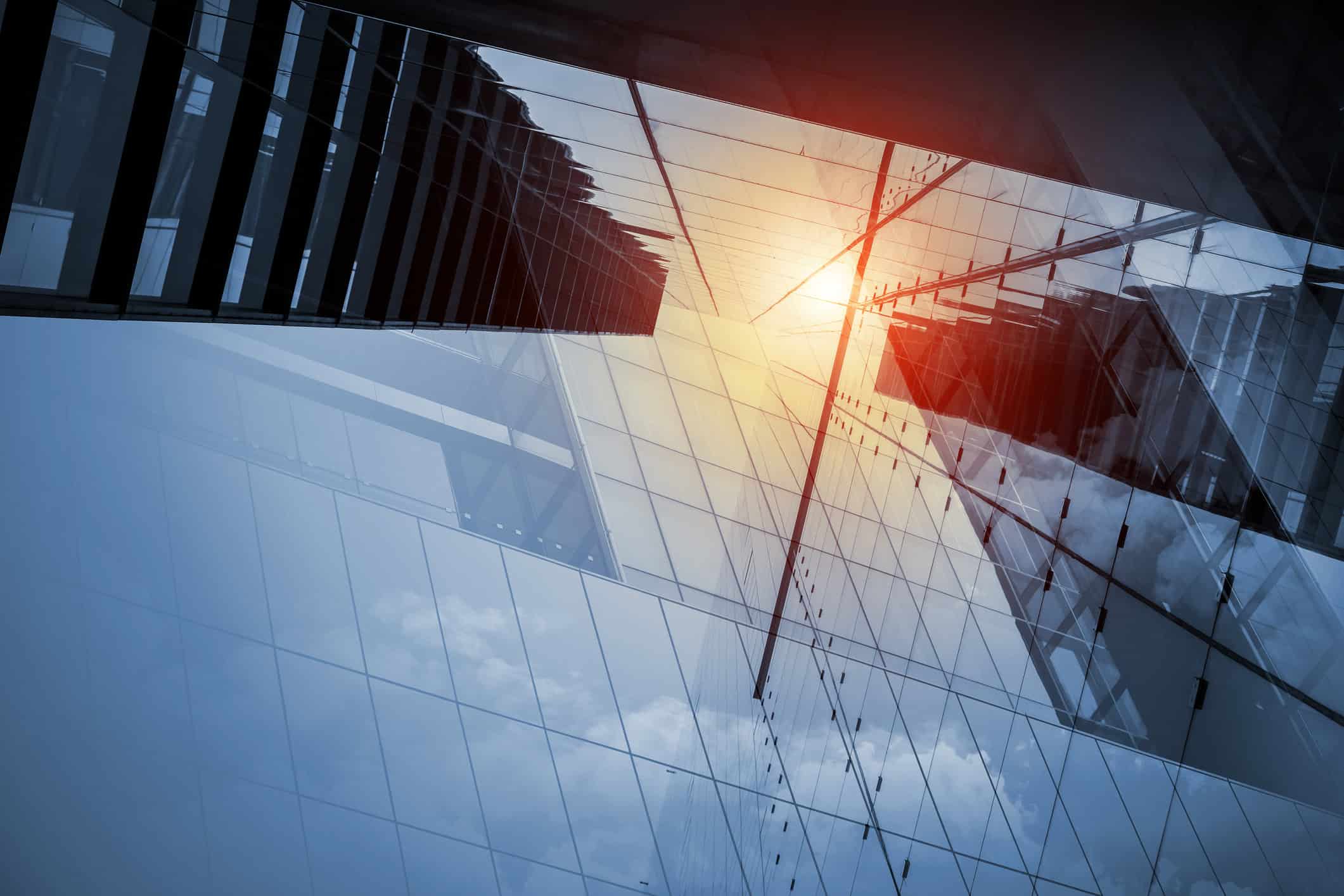 Low-angle view of a modern glass building facade with reflections of other structures and sky, featuring a bright orange and red sun flare.