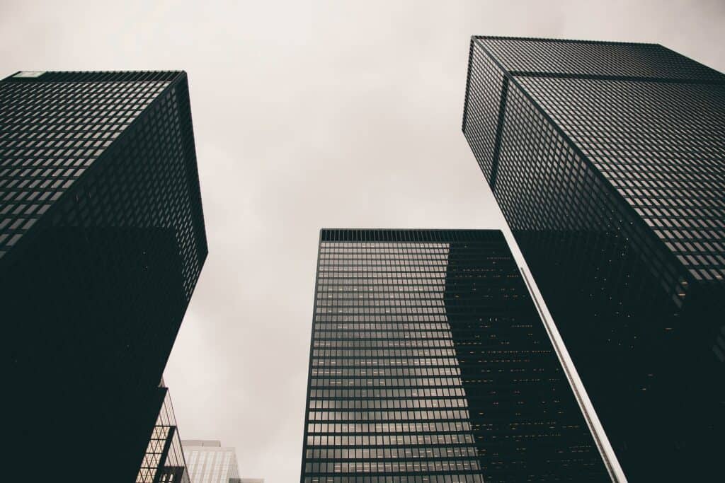 Three dark, modern skyscrapers seen from a low angle against a light gray, overcast sky. The buildings feature grid-patterned windows.