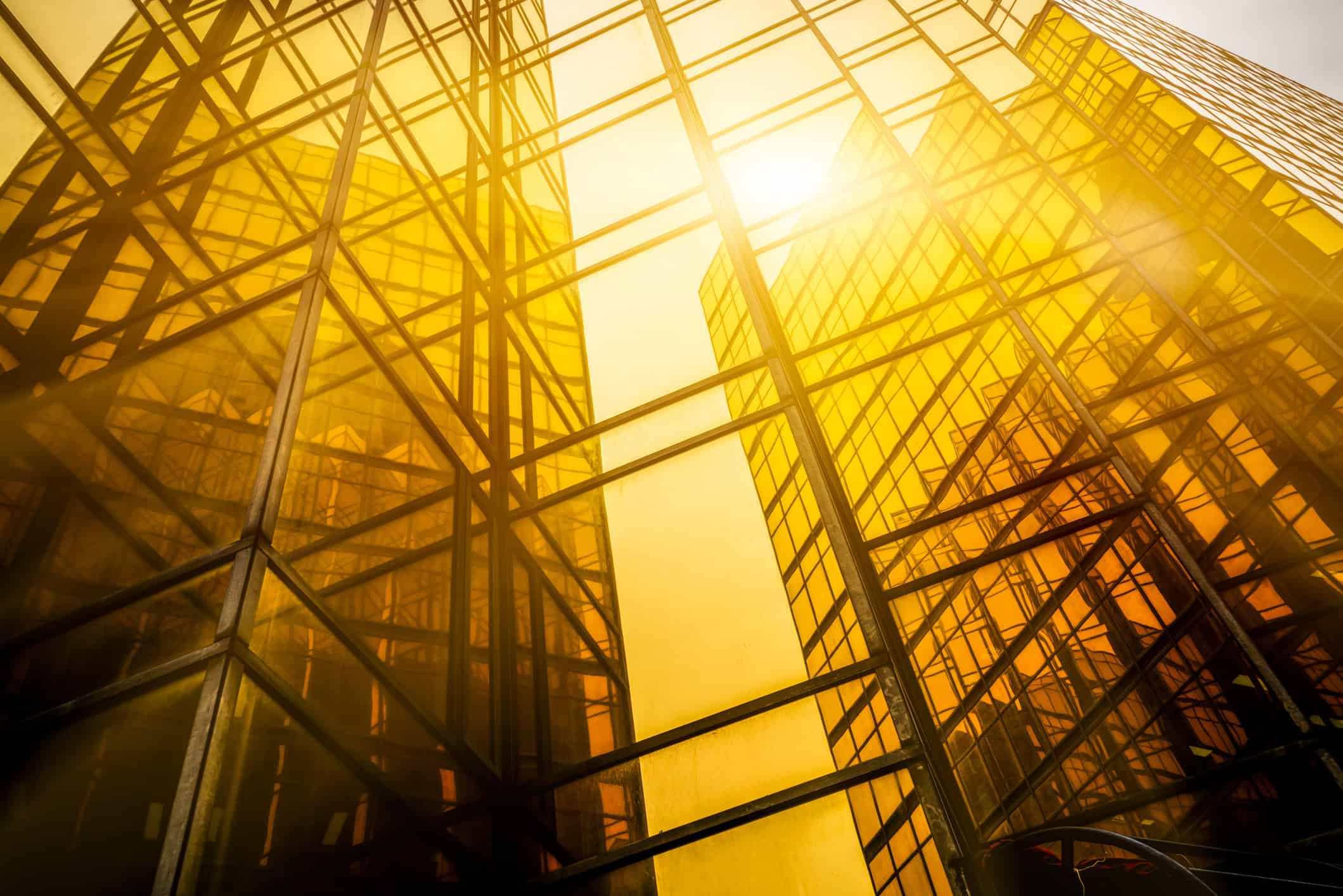 Low-angle view of a modern building with a golden glass facade and dark grid frames, brightly lit by a sun flare.