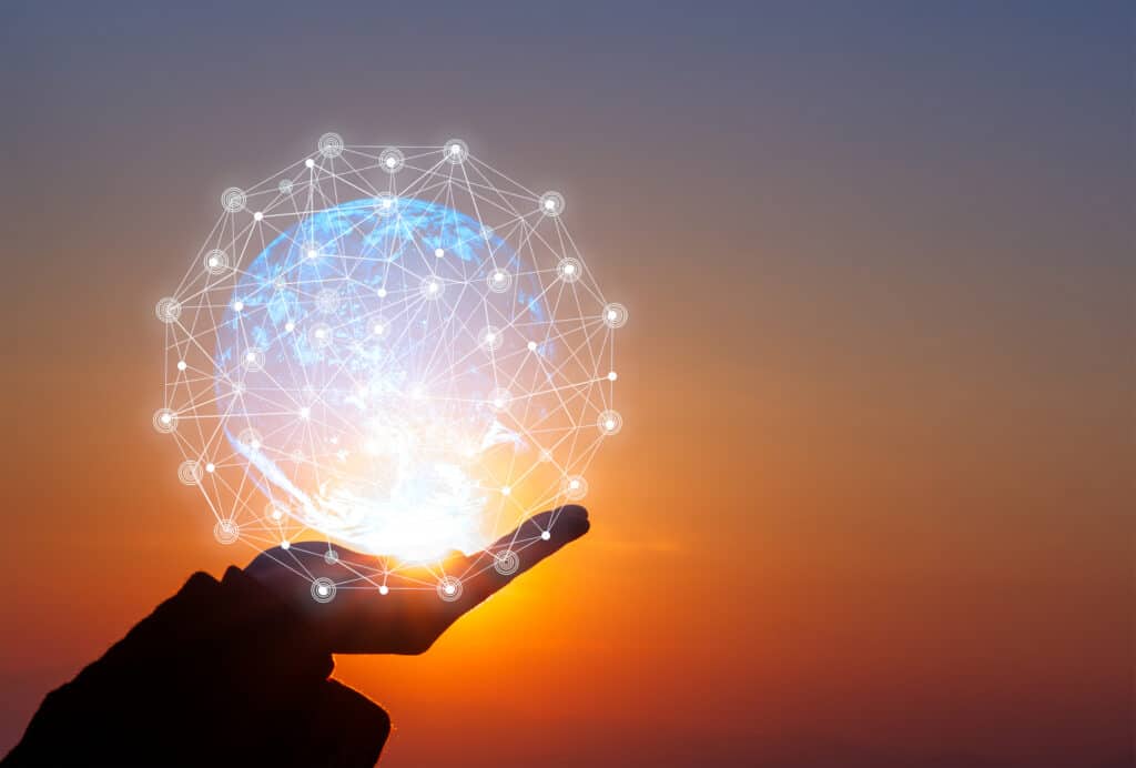 Silhouette of a hand holding a glowing blue digital globe with a white network overlay against an orange and purple sunset sky.