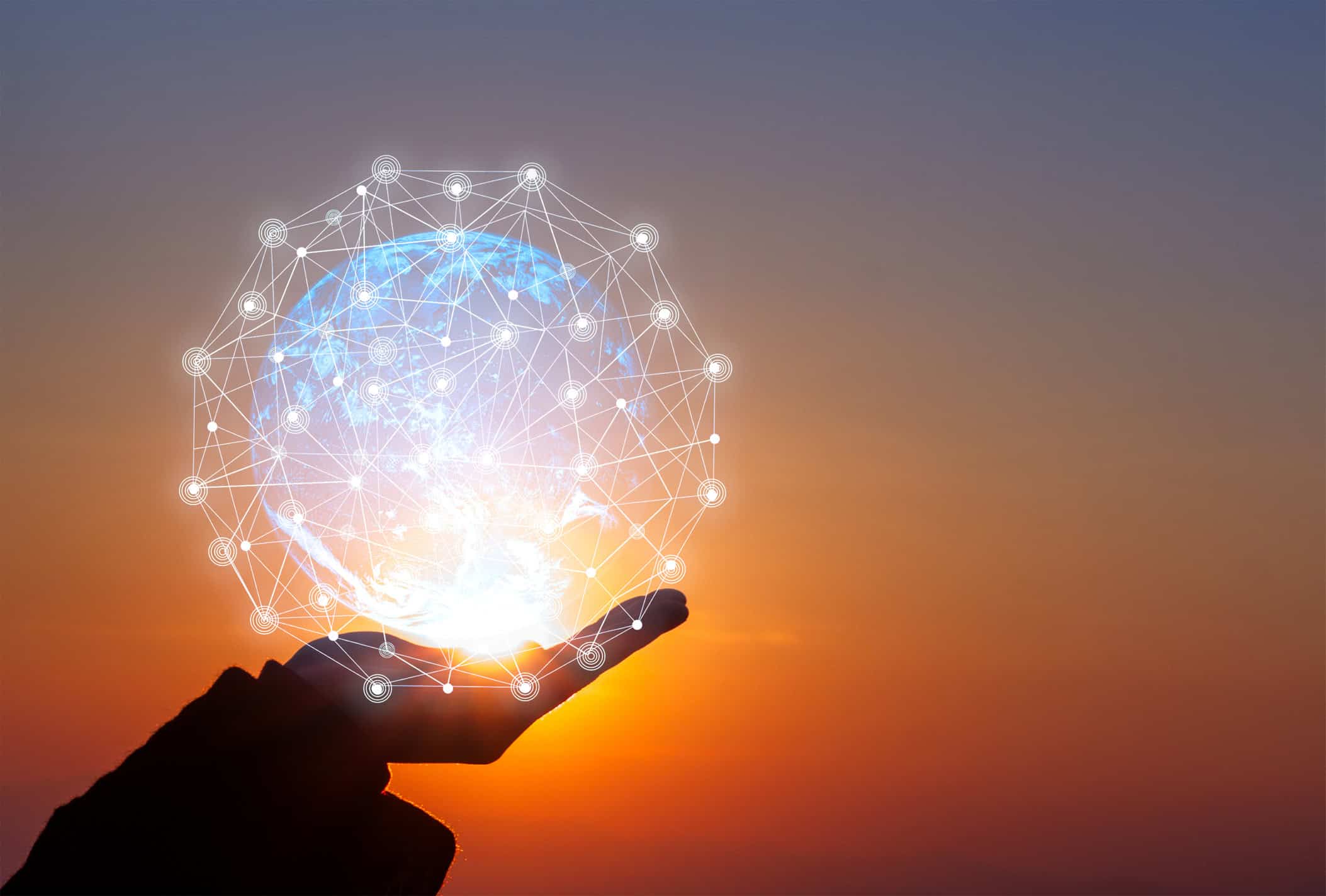 Silhouette of a hand holding a glowing blue digital globe with a white network overlay against an orange and purple sunset sky.