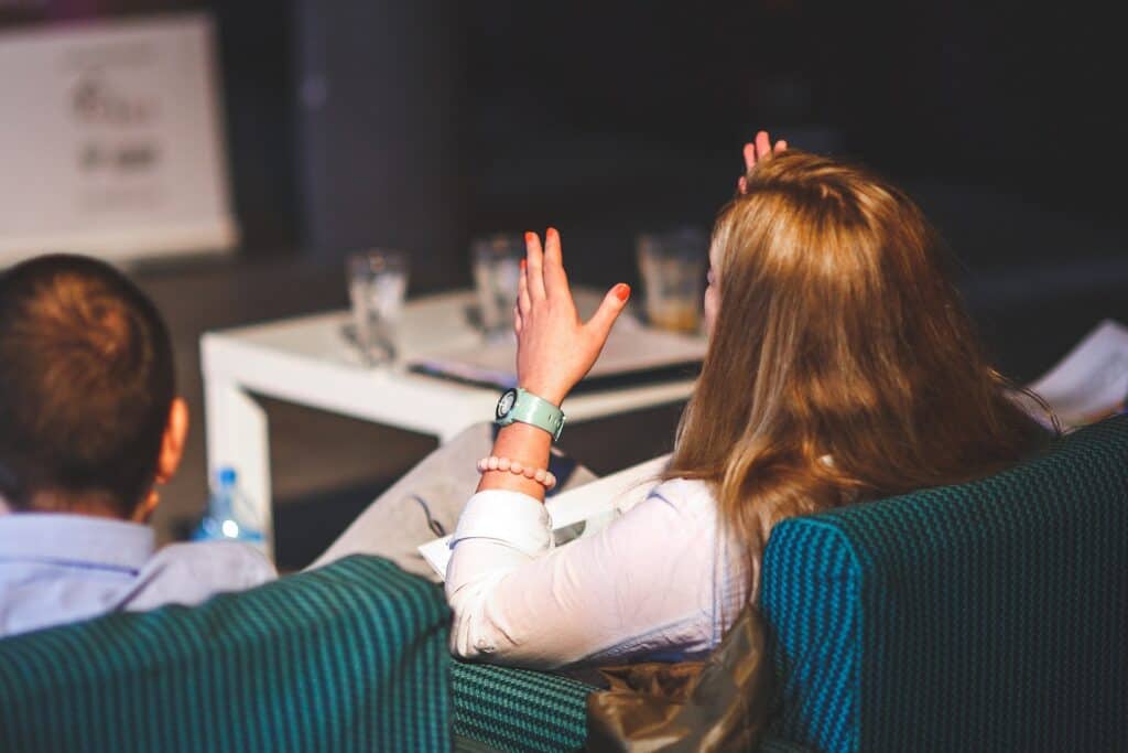 Rear view of a woman with long hair raising her hand, seated in a conference with another person and a table with drinks.