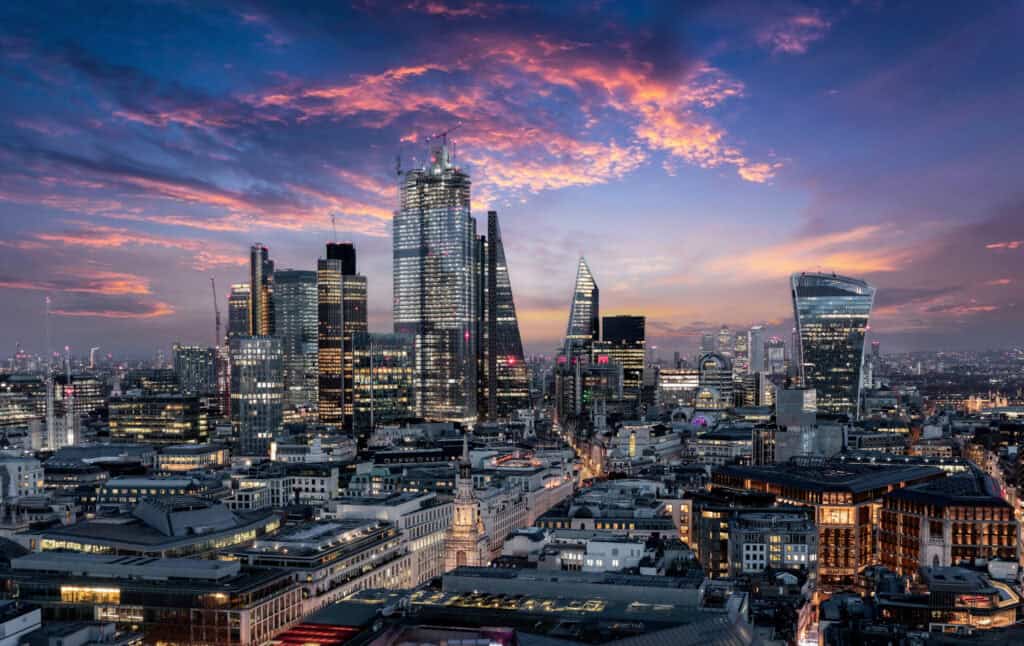 Aerial view of London's cityscape at dusk, featuring illuminated skyscrapers and smaller buildings under a dramatic sky with pink, orange, and blue clouds.