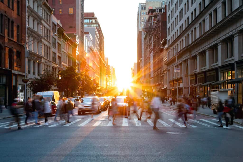 A city street at sunset, with bright sun at the end of the street, blurring pedestrians and cars in motion.