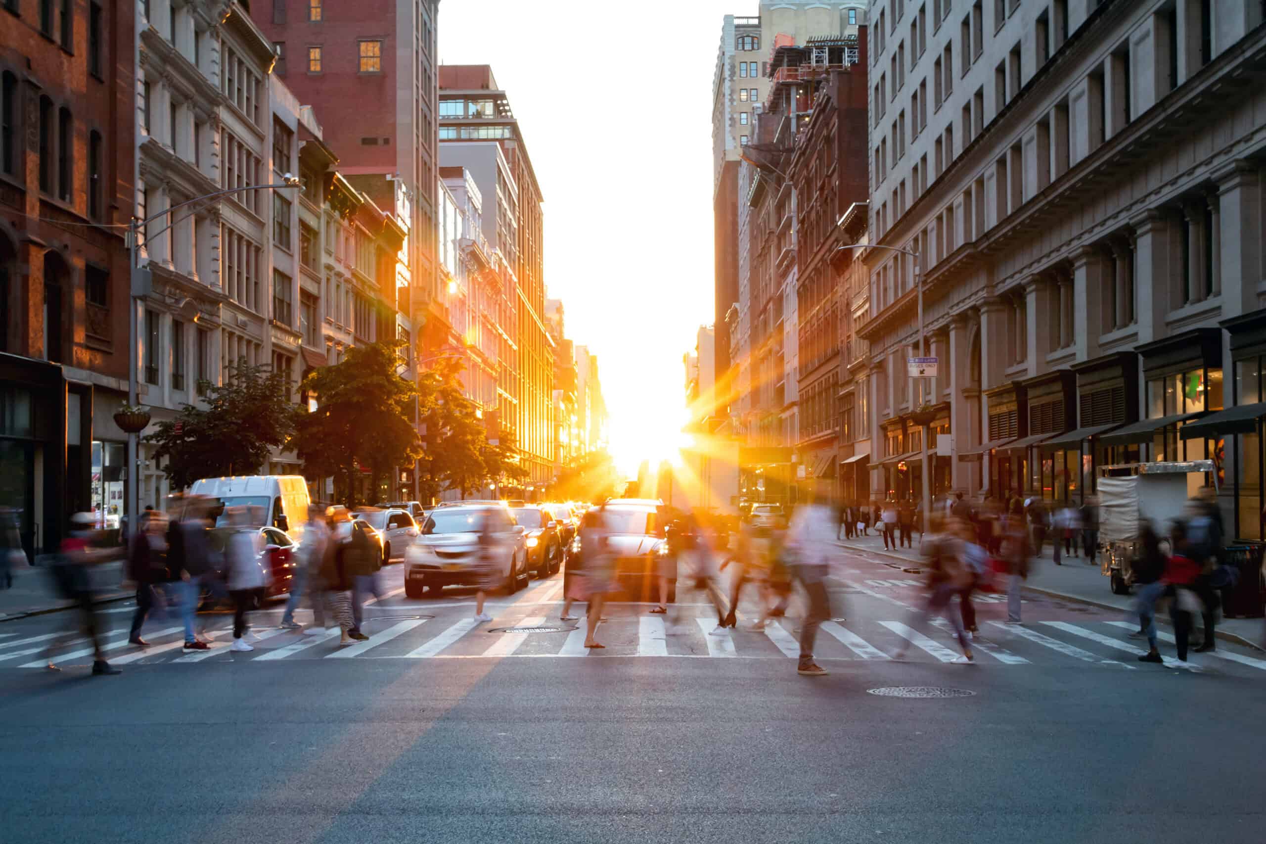 A city street at sunset, with bright sun at the end of the street, blurring pedestrians and cars in motion.