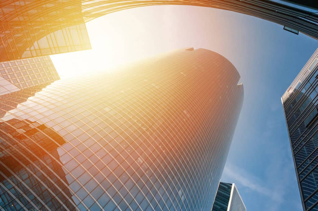 Low-angle view of tall, curved glass skyscrapers under a blue sky with a bright sun flare.