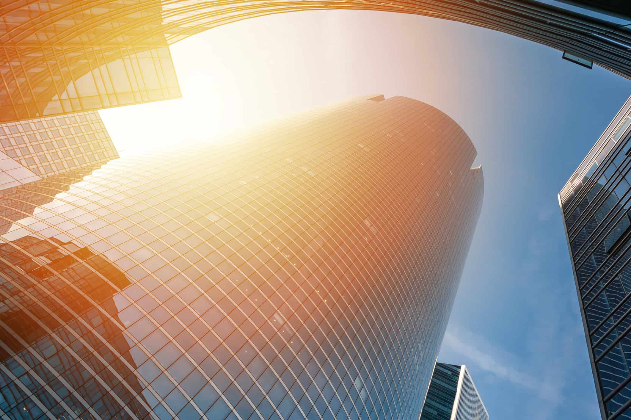 Low-angle view of tall, curved glass skyscrapers under a blue sky with a bright sun flare.