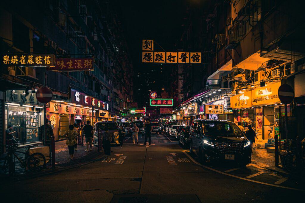 Nighttime street scene in an Asian city with tall buildings, numerous illuminated Chinese signs, pedestrians, and cars.