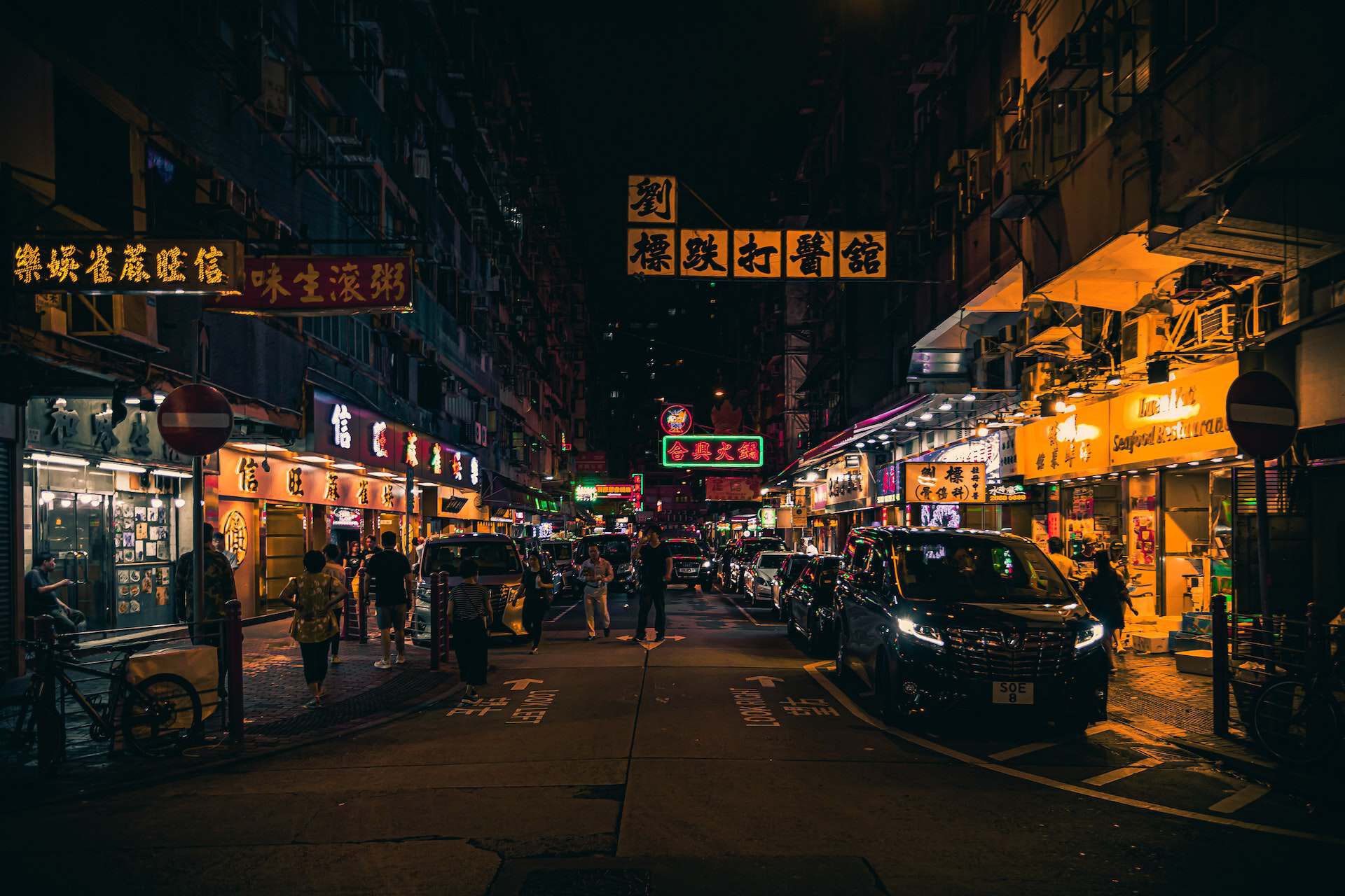 Nighttime street scene in an Asian city with tall buildings, numerous illuminated Chinese signs, pedestrians, and cars.