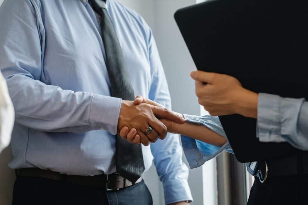 Two people in business attire shaking hands, one wearing a light blue shirt and tie, the other holding a black folder.