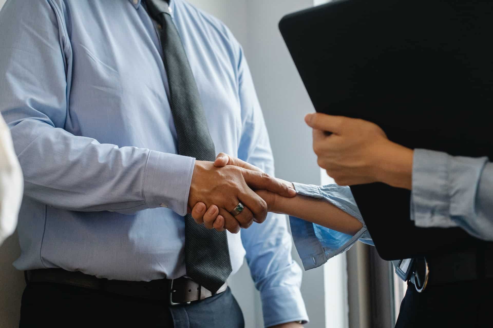 Two people in business attire shaking hands, one wearing a light blue shirt and tie, the other holding a black folder.