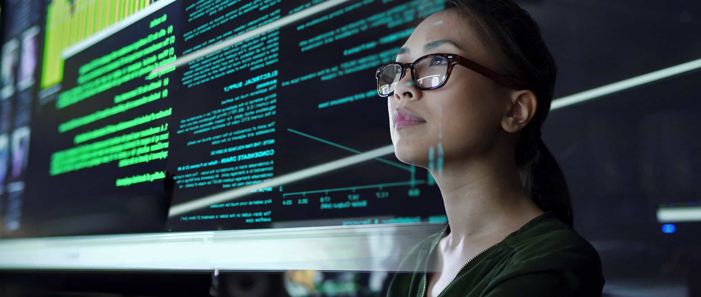 Side profile of a woman in glasses looking at multiple large screens displaying green code and graphs.