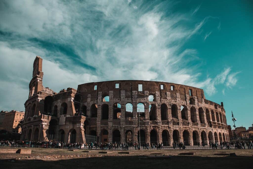 The Colosseum in Rome, a large, ancient amphitheater made of stone and brick, with numerous arches and partially ruined sections, under a dramatic teal-blue sky with white clouds. A crowd of people gathers at its base.