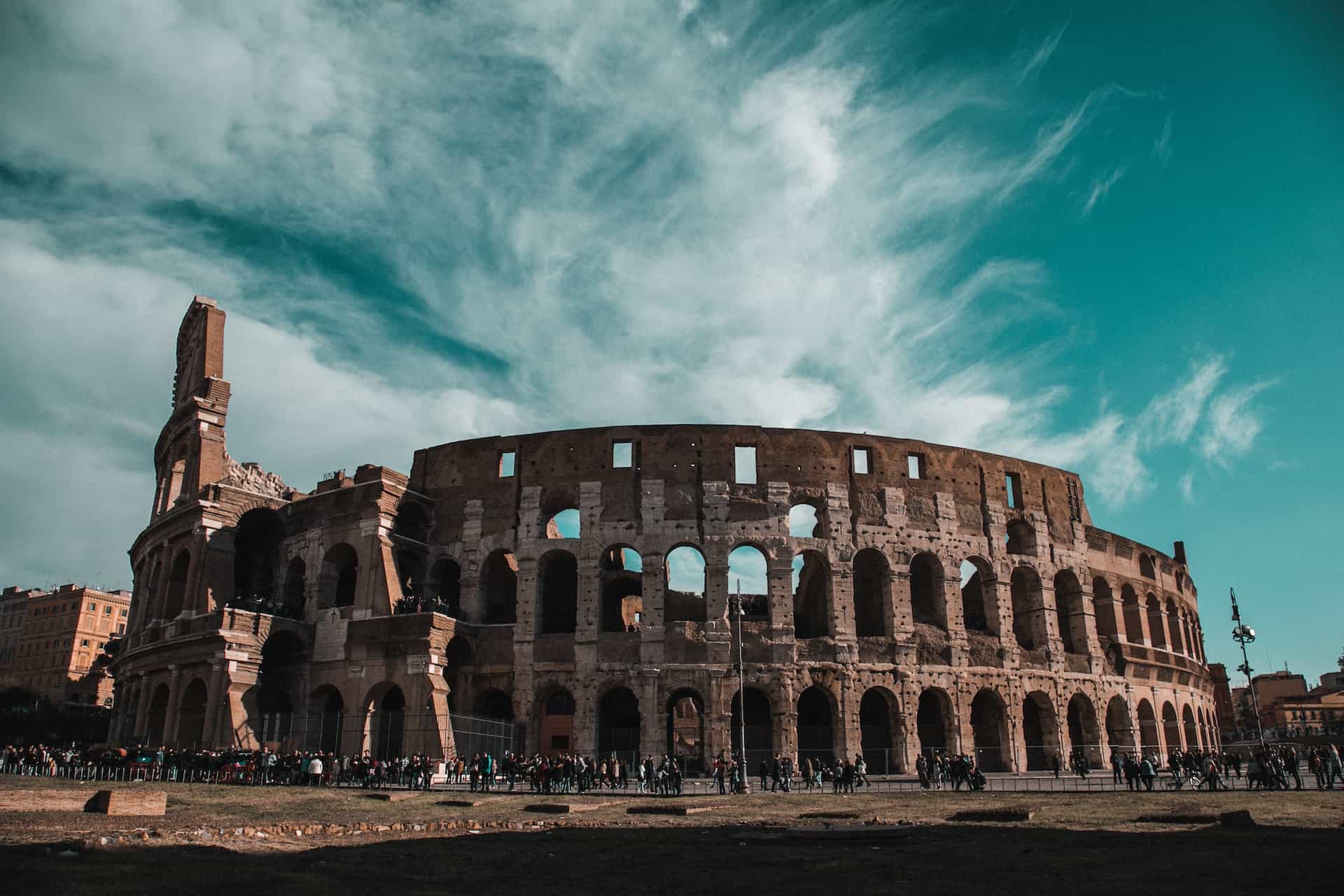 The Colosseum in Rome, a large, ancient amphitheater made of stone and brick, with numerous arches and partially ruined sections, under a dramatic teal-blue sky with white clouds. A crowd of people gathers at its base.