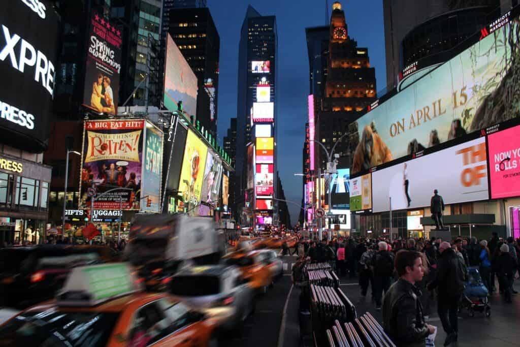 Times Square at night, with bright billboards, blurred traffic, and a crowd of people on the sidewalks.