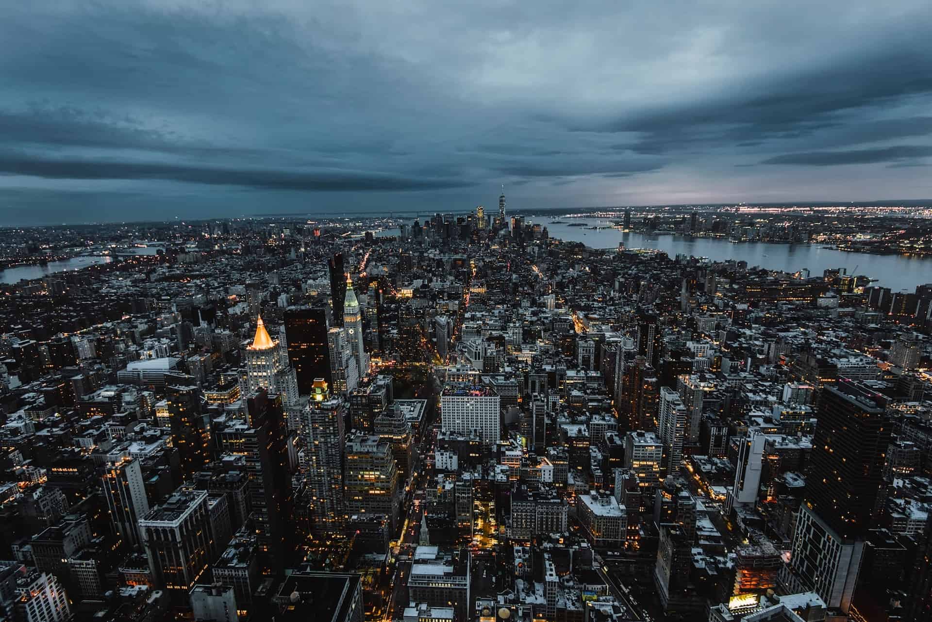 Aerial view of New York City at dusk, showing a vast urban landscape with thousands of illuminated buildings, including One World Trade Center and the Chrysler Building, under a dark, cloudy sky. Rivers flank the city.