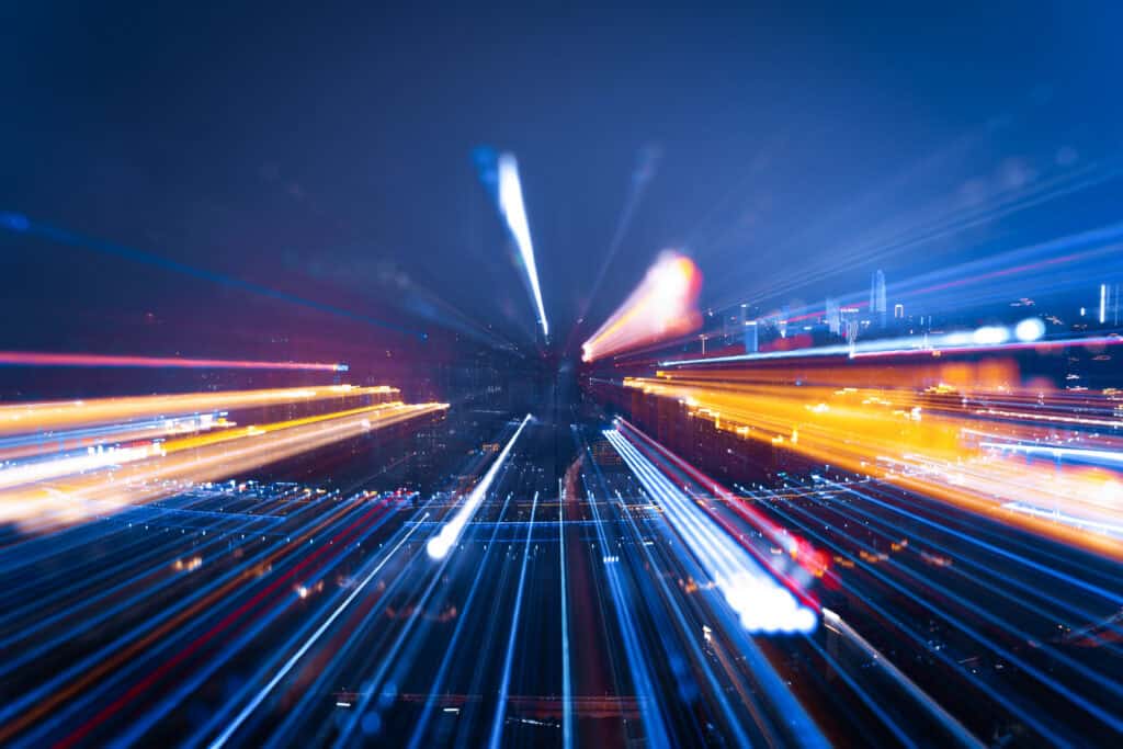 Abstract image of a city at night with long, colorful light trails in blue, orange, red, and white, creating a dynamic motion blur effect.