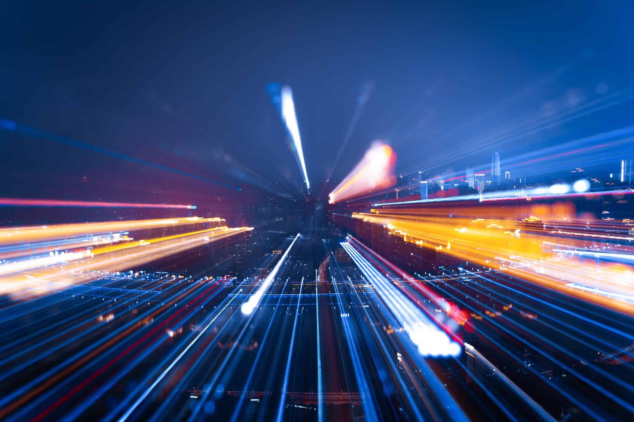 Abstract image of a city at night with long, colorful light trails in blue, orange, red, and white, creating a dynamic motion blur effect.