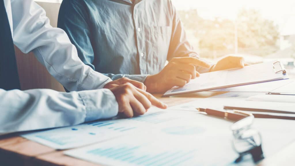 Two people reviewing charts and graphs on documents and a clipboard at a table, with one person pointing at the data.