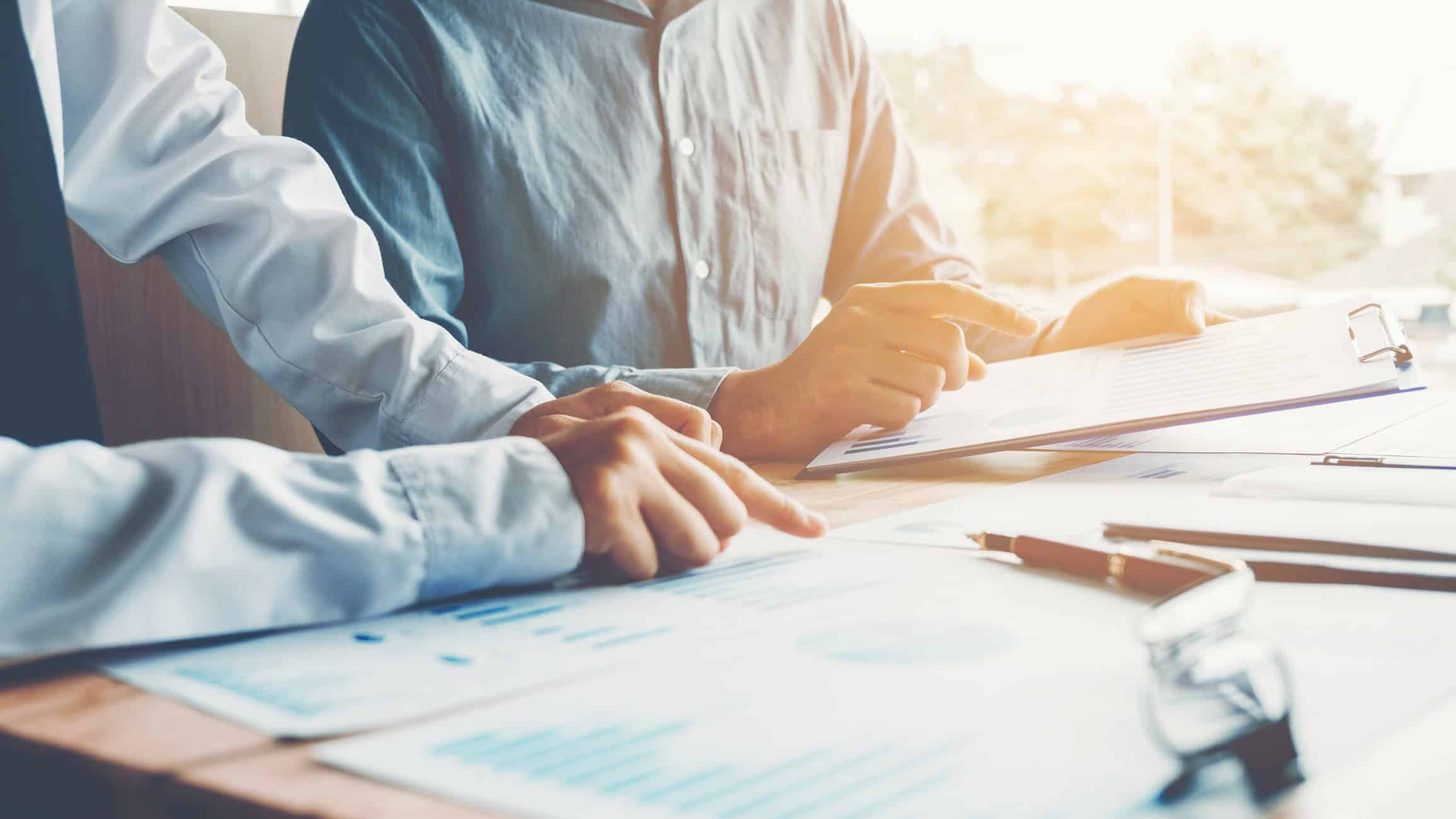 Two people reviewing charts and graphs on documents and a clipboard at a table, with one person pointing at the data.