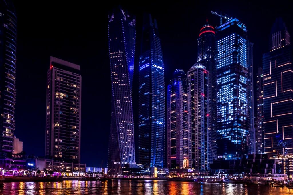Night view of a cityscape with numerous illuminated skyscrapers reflecting on a body of water.