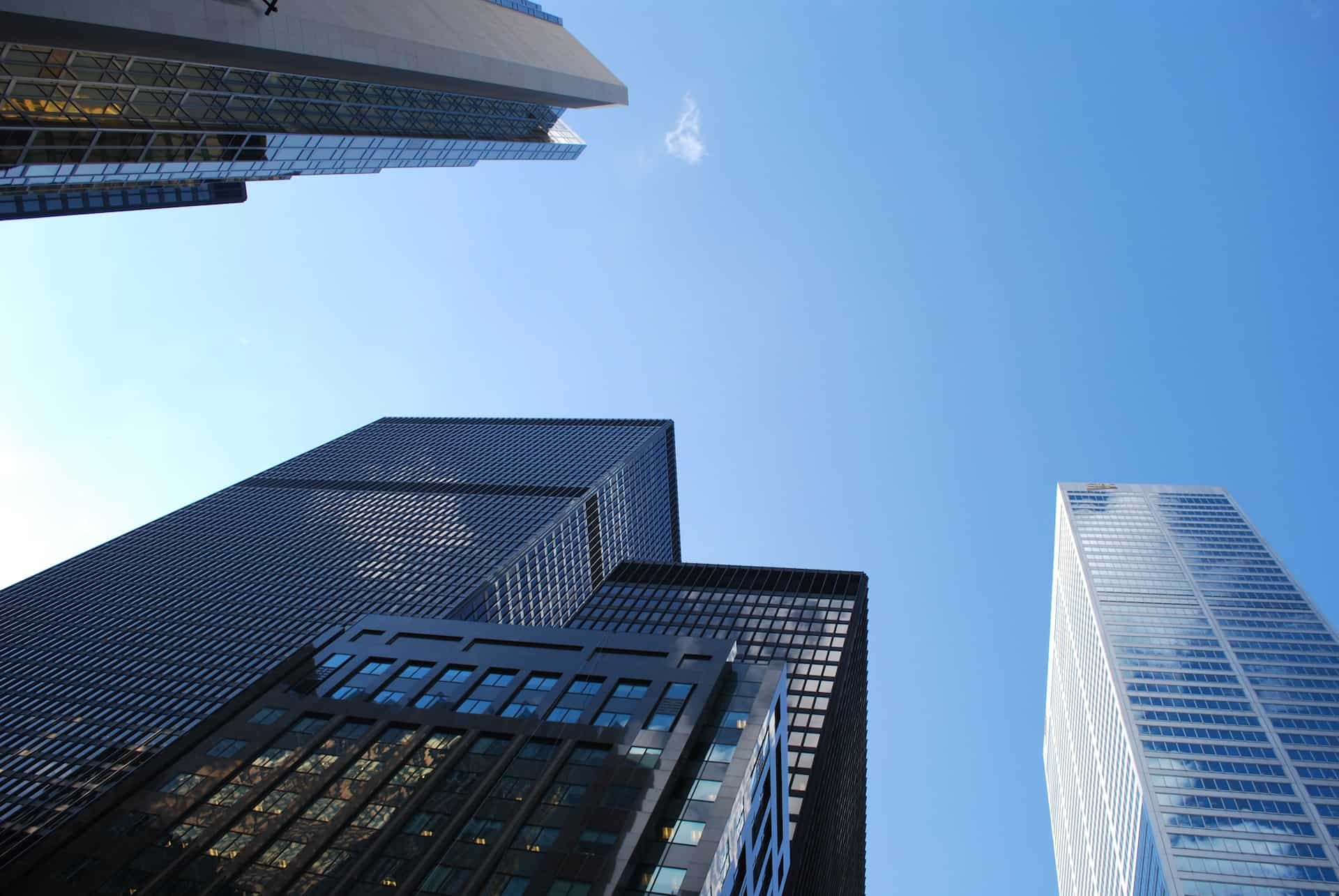 A worm's-eye view of multiple modern skyscrapers with glass and dark facades against a clear blue sky.