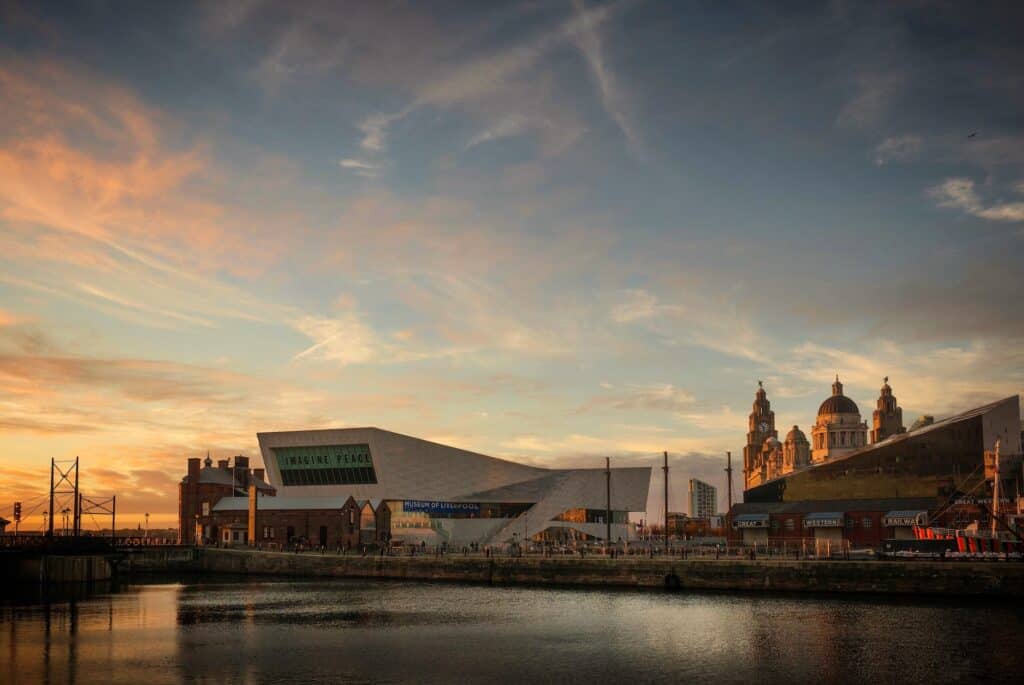 Liverpool waterfront at sunset, showing the modern Museum of Liverpool alongside historic buildings with domes and towers, reflected in dark water under an orange and blue sky.