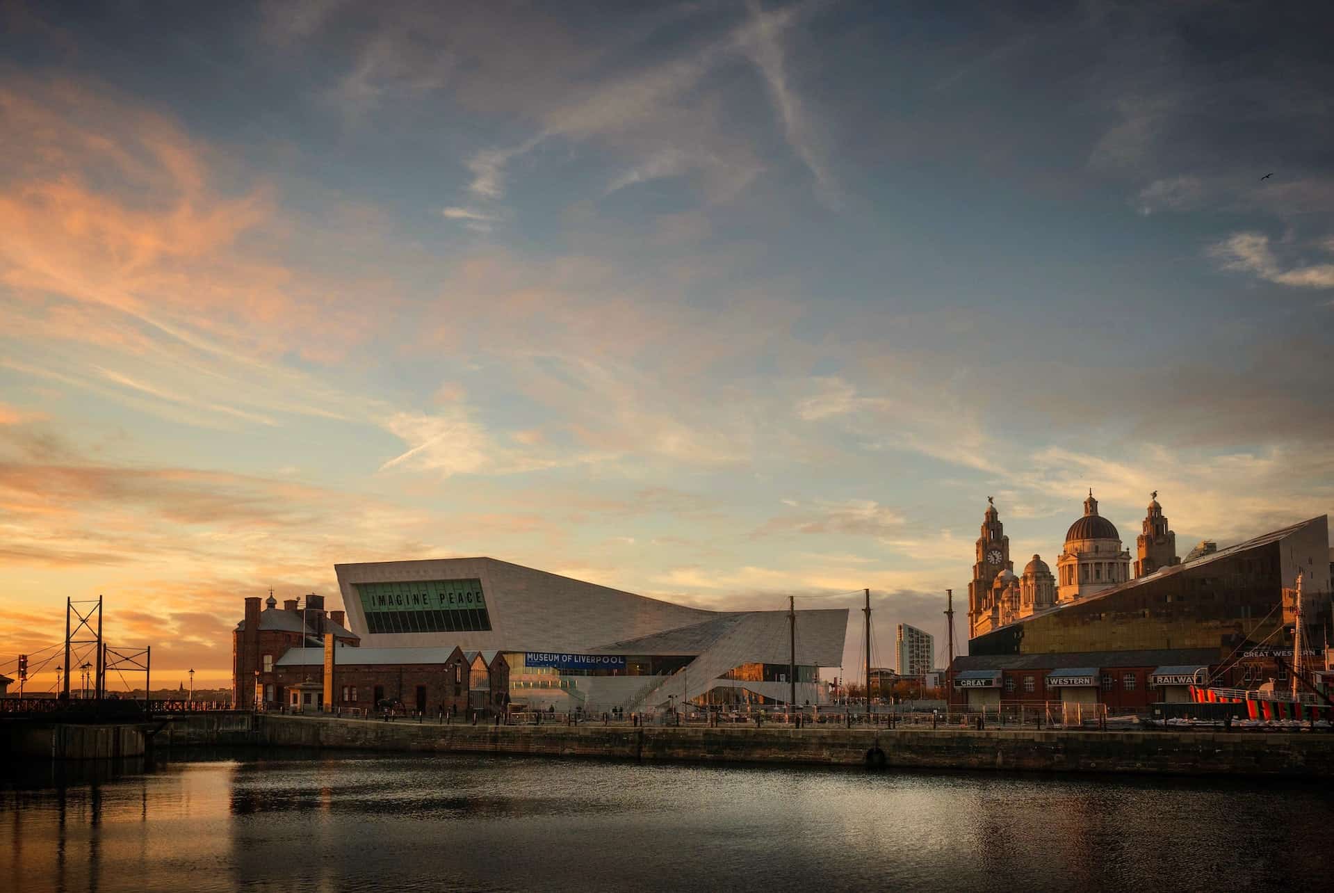 Liverpool waterfront at sunset, showing the modern Museum of Liverpool alongside historic buildings with domes and towers, reflected in dark water under an orange and blue sky.