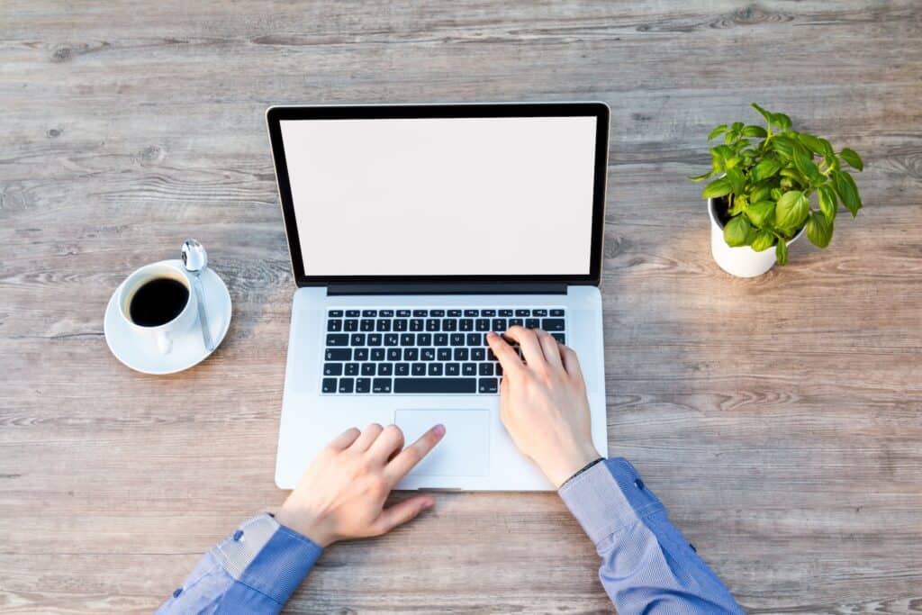 Overhead view of hands typing on a laptop with a blank screen, a cup of coffee, and a potted plant on a wooden desk.