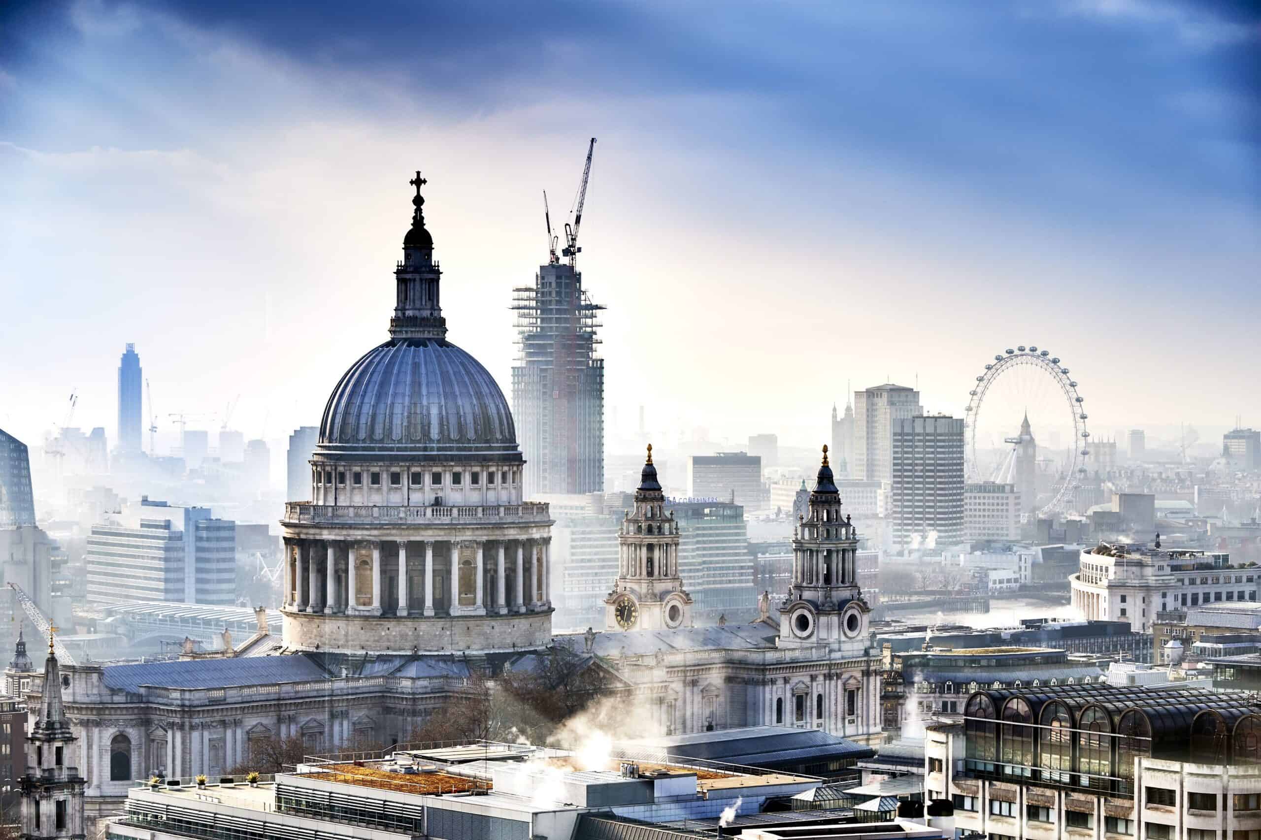 Aerial view of St. Paul's Cathedral with its dome and towers, surrounded by a hazy London cityscape including a skyscraper under construction and the London Eye in the distance.