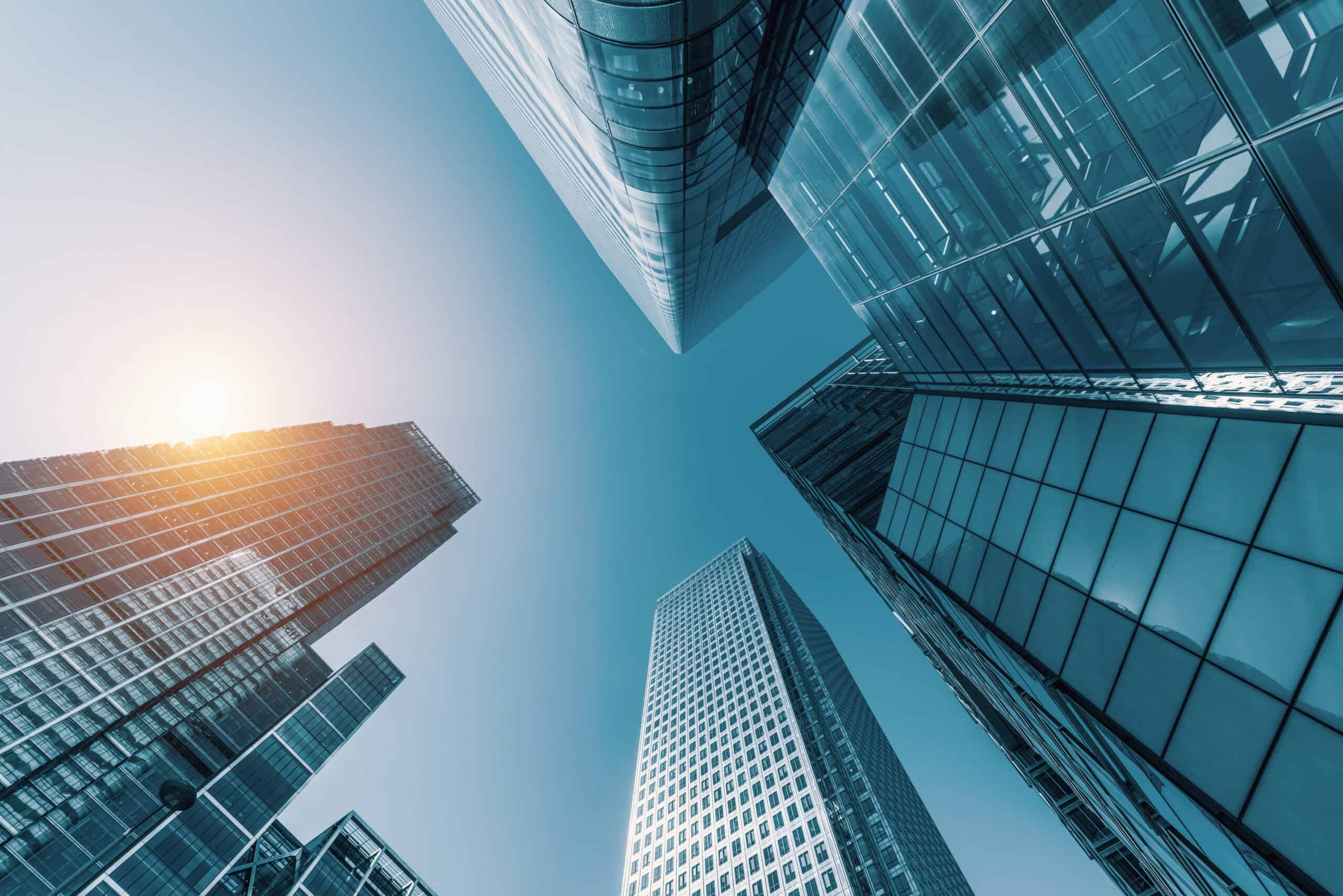 Low-angle view of multiple modern glass skyscrapers against a clear blue sky with a sun flare.