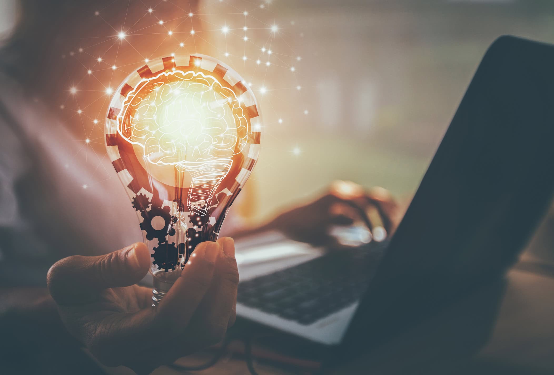 A hand holds a glowing lightbulb containing a brain graphic and gears, with network lines emanating from it. A person types on a laptop in the blurry background.