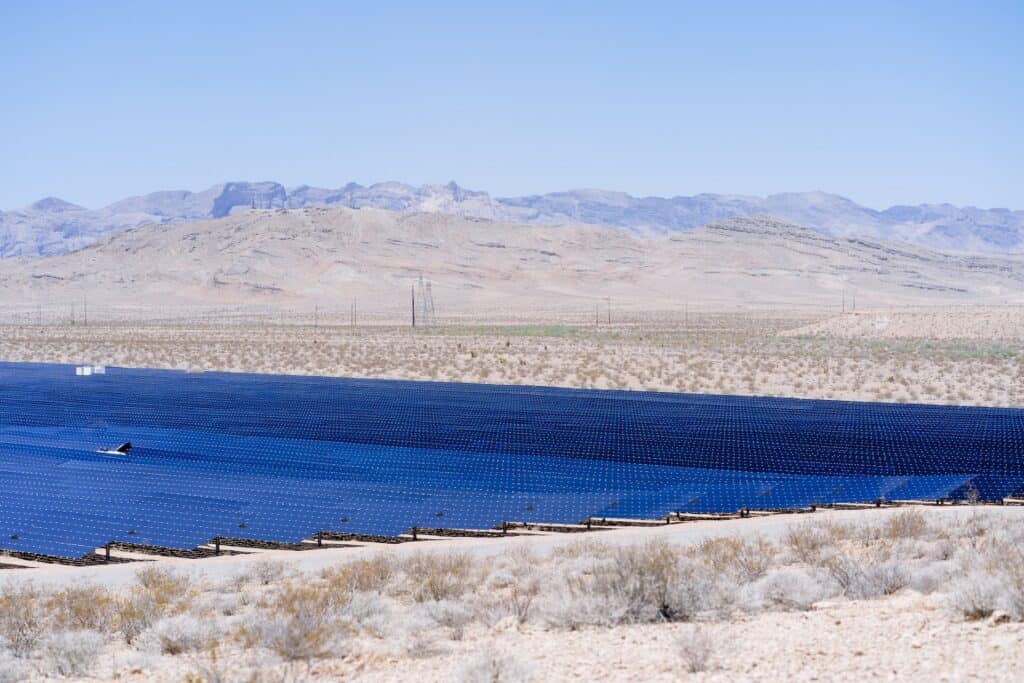 A large solar farm with rows of dark blue panels in a desert landscape, with mountains and power lines in the background under a clear sky.