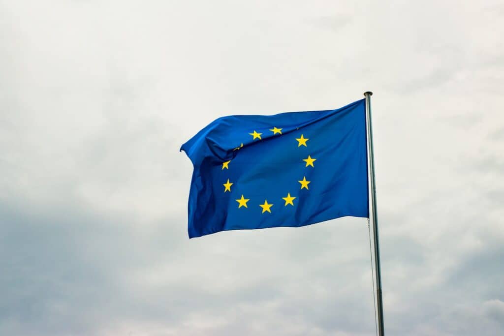 The European Union flag, blue with a circle of twelve golden stars, waves from a flagpole against a cloudy sky.