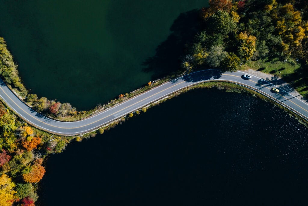 Aerial view of a winding road between two dark lakes, surrounded by trees with red, orange, and yellow autumn leaves. Two cars are visible on the road.