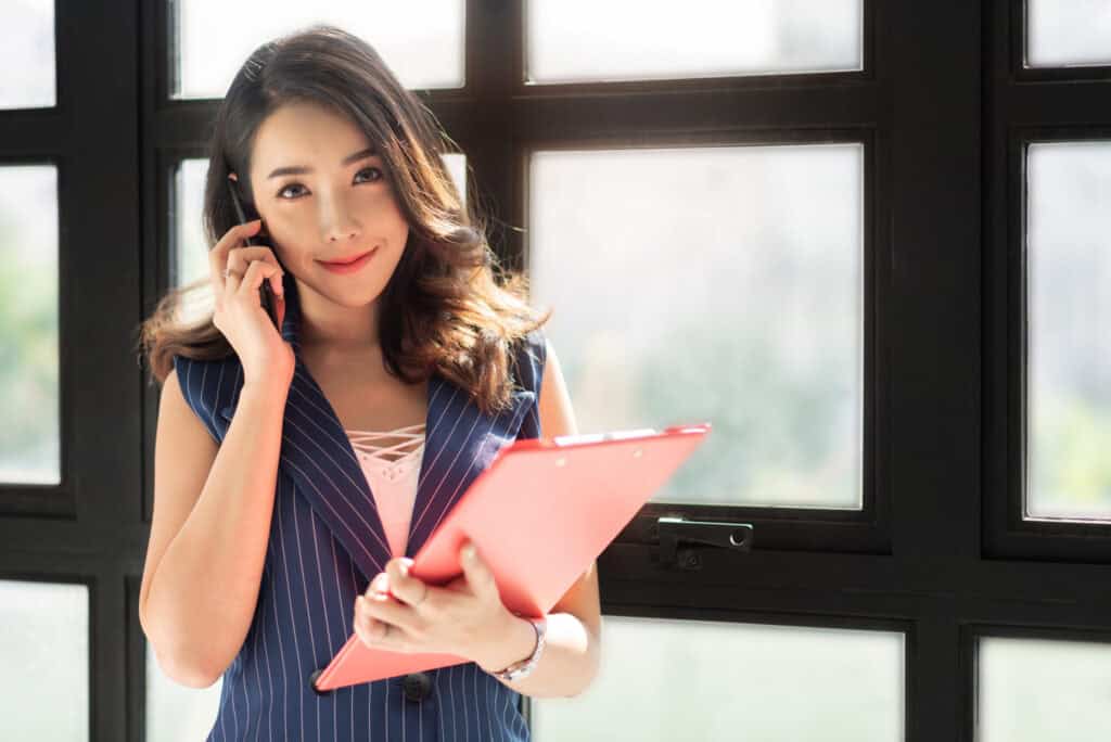 A young Asian woman on a phone call, holding a red clipboard, standing by a large window.