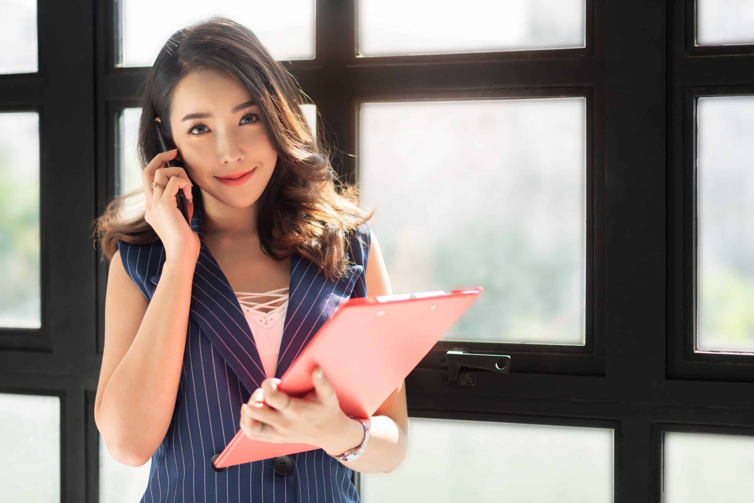 A young Asian woman on a phone call, holding a red clipboard, standing by a large window.