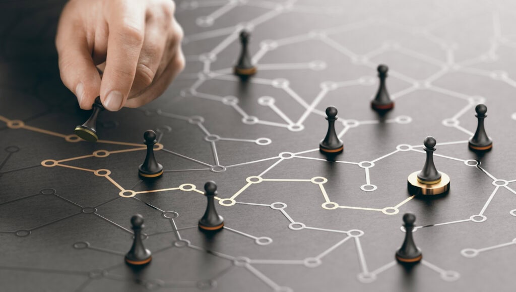A hand moves a black chess pawn with a gold base on a dark, textured surface featuring a network grid of interconnected white and gold lines and circles. Other black pawns are scattered across the grid.